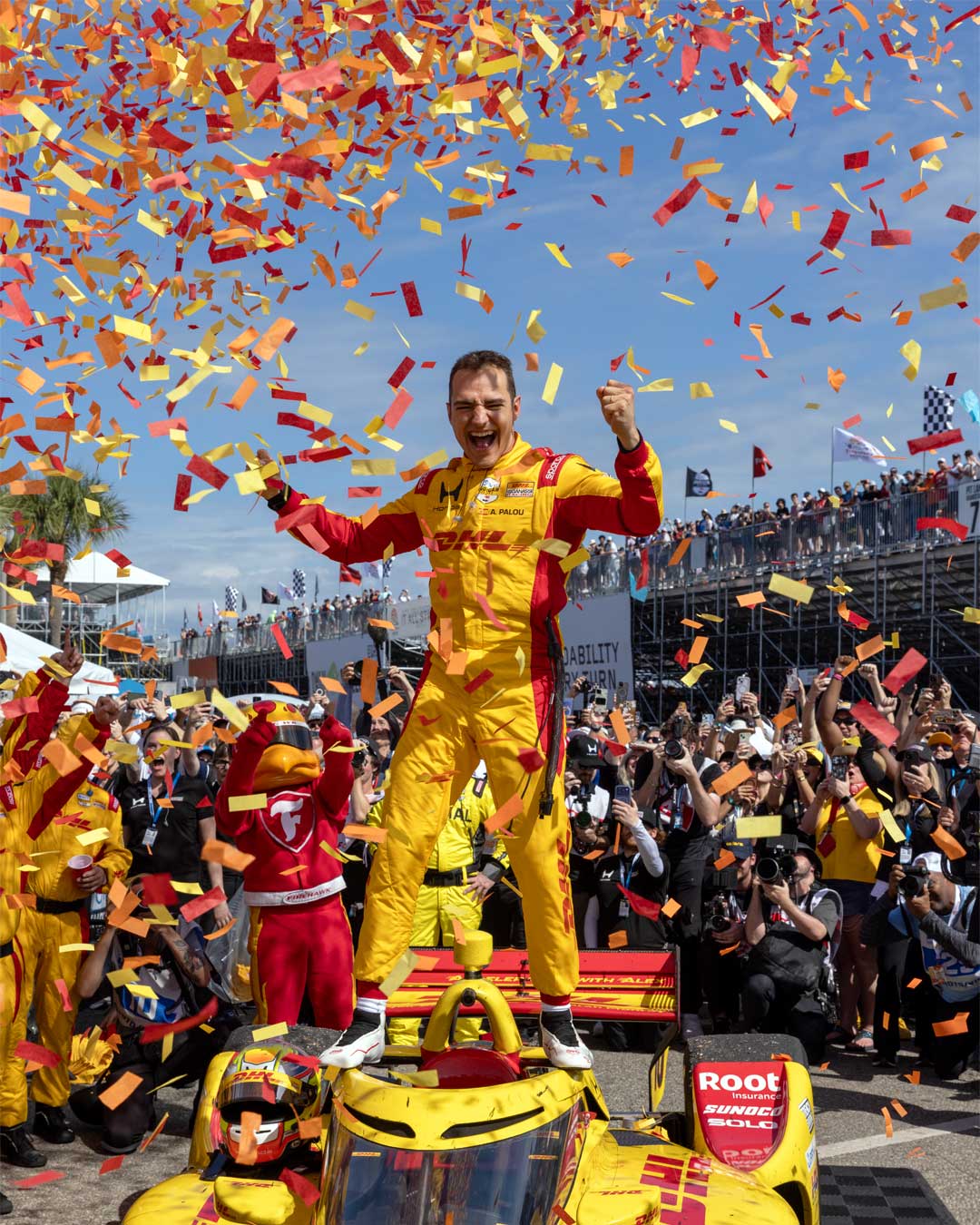 Alex Palou stands on his car with arms raised as orange and yellow confetti fills the air